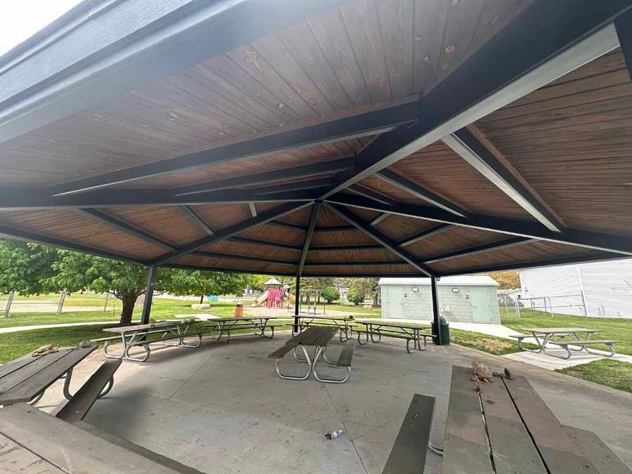 Covered outdoor pavilion with picnic tables and benches on a concrete floor, viewed from underneath the roof