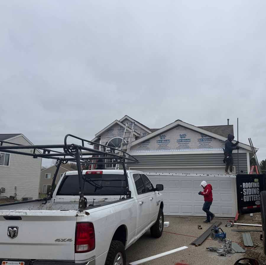 White pickup truck in front of a storm-damaged building, with a worker on a ladder and debris on the ground.
