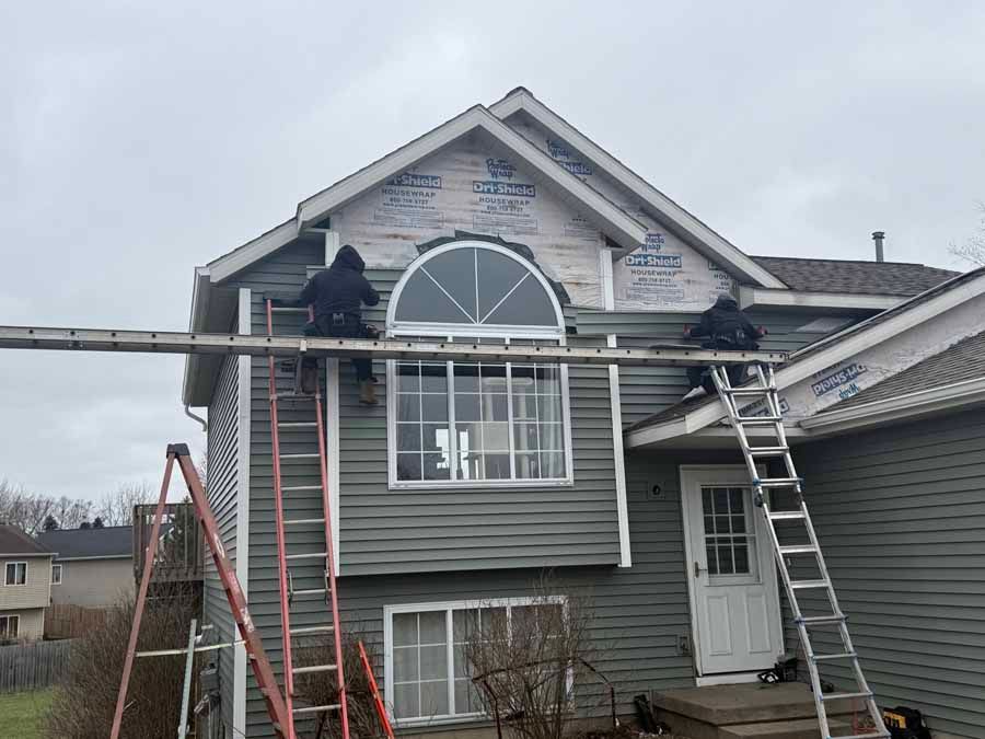 Gray two-story house under repair with ladders, scaffolding, and a person on the roof