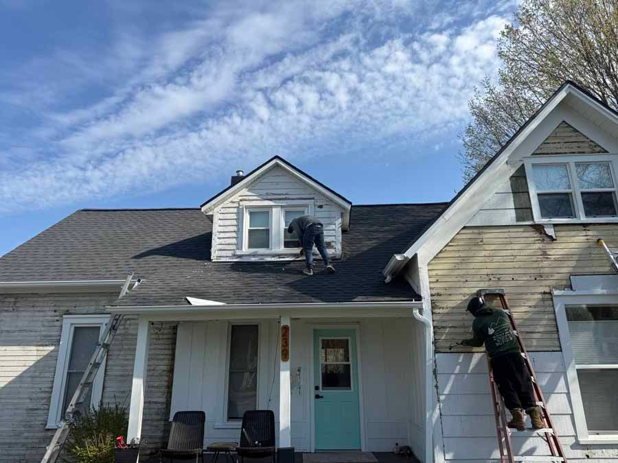 Workers on a house roof and ladder, cleaning gutters on a light-colored home under a blue sky.