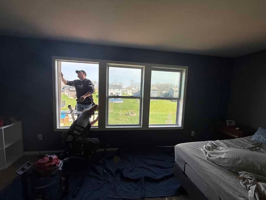 Person on a ladder working on the window in a dark bedroom with an unmade bed and sunlight outside.