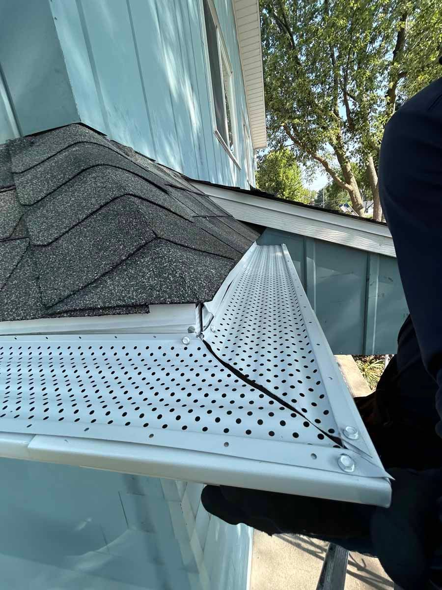 Person holding a white metal roof vent next to a house with gray shingles and blue siding