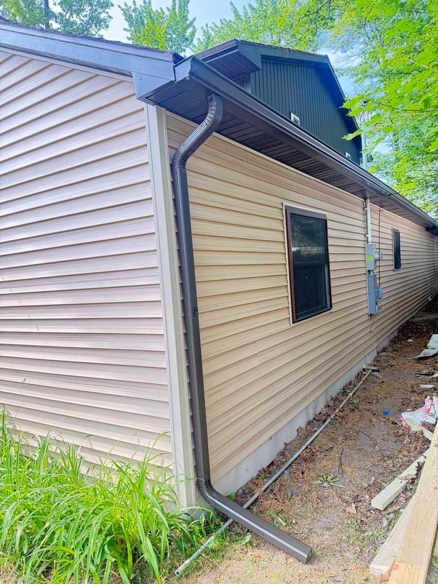 Side view of a beige house with a damaged roof, gutter downspout, and trees in the background