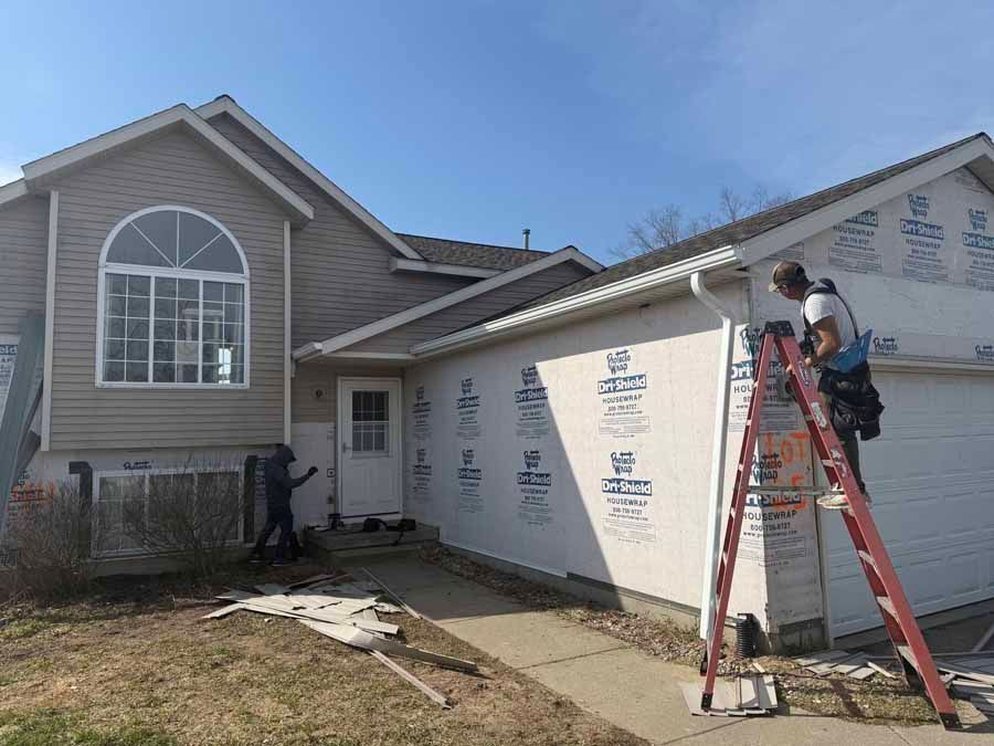 Two workers installing white house siding, one on a ladder beside a gray home with a large window.