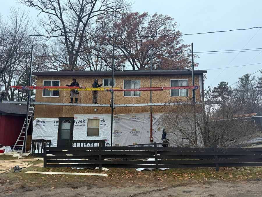 Two-story house under renovation with workers on an upper balcony and scaffolding in front