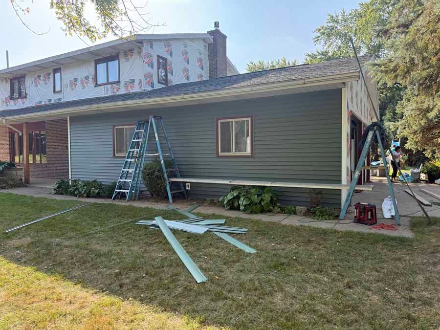 Workers installing siding and trim on a house exterior, with ladders and materials on the lawn.