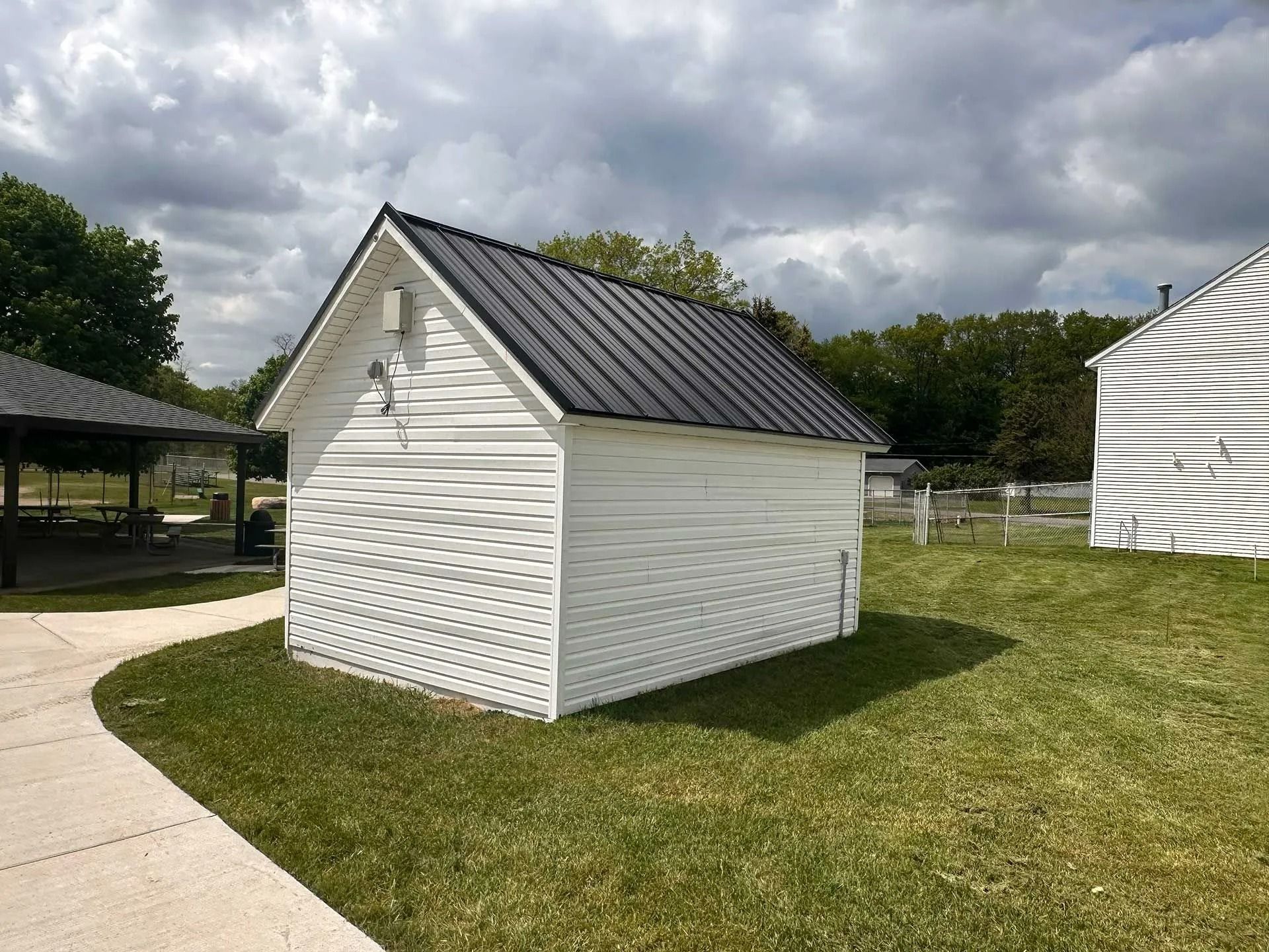 Small white shed with a dark metal roof beside a curved sidewalk on a grassy lawn.