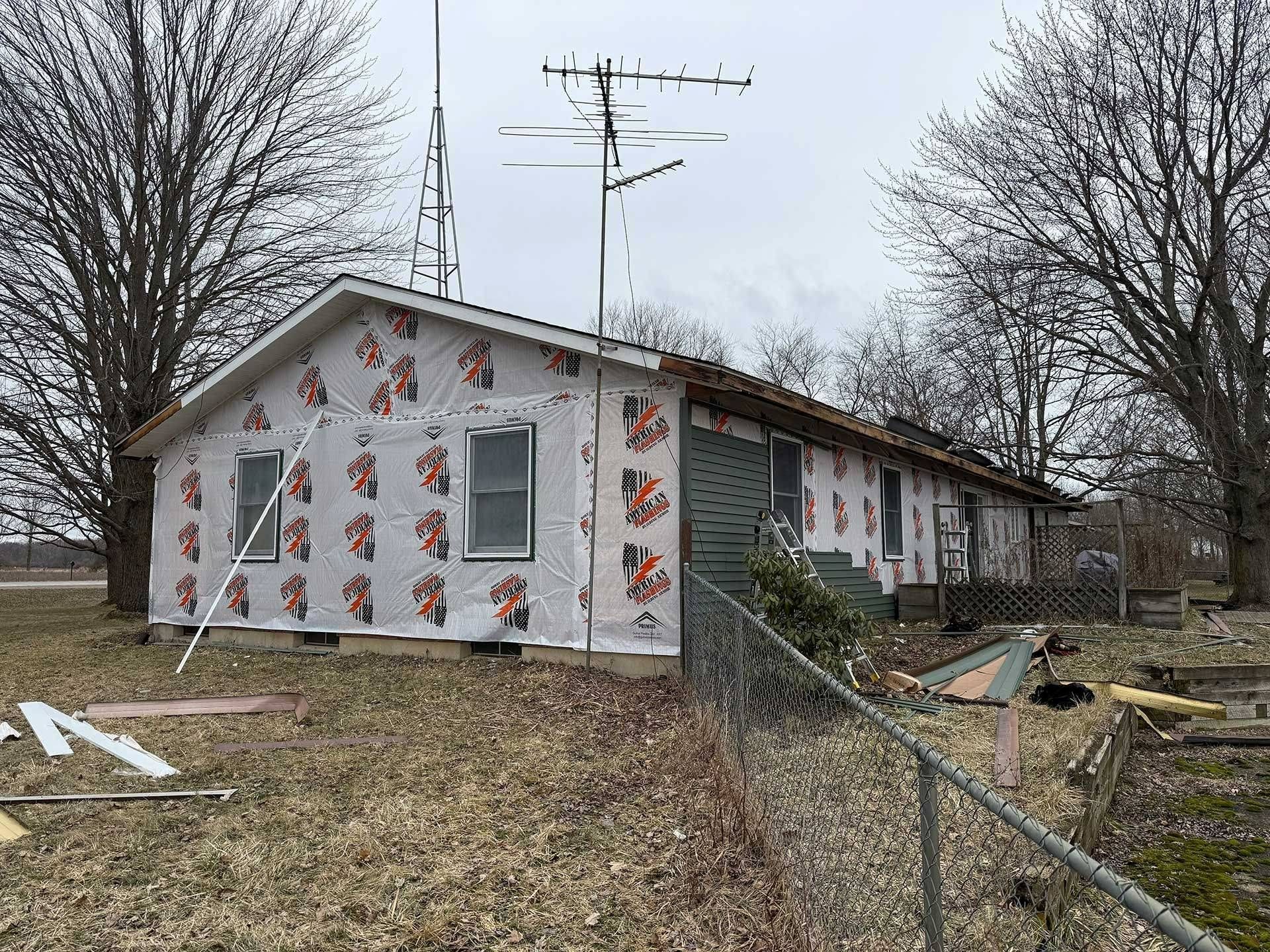 Small house under repair with exposed insulation, chain-link fence, and bare trees in a yard