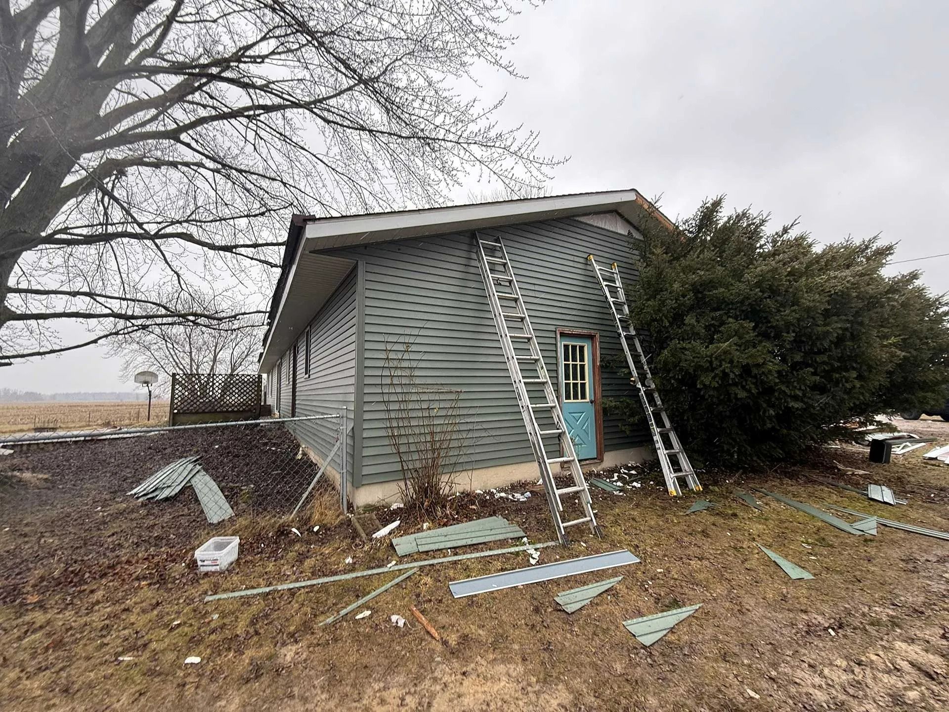 Damaged gray house with shattered windows, debris on the ground, and a ladder leaning against the side.