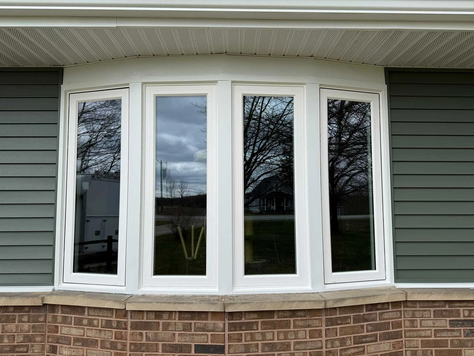 White bay window in green siding and brick house exterior with curtains and tree reflections