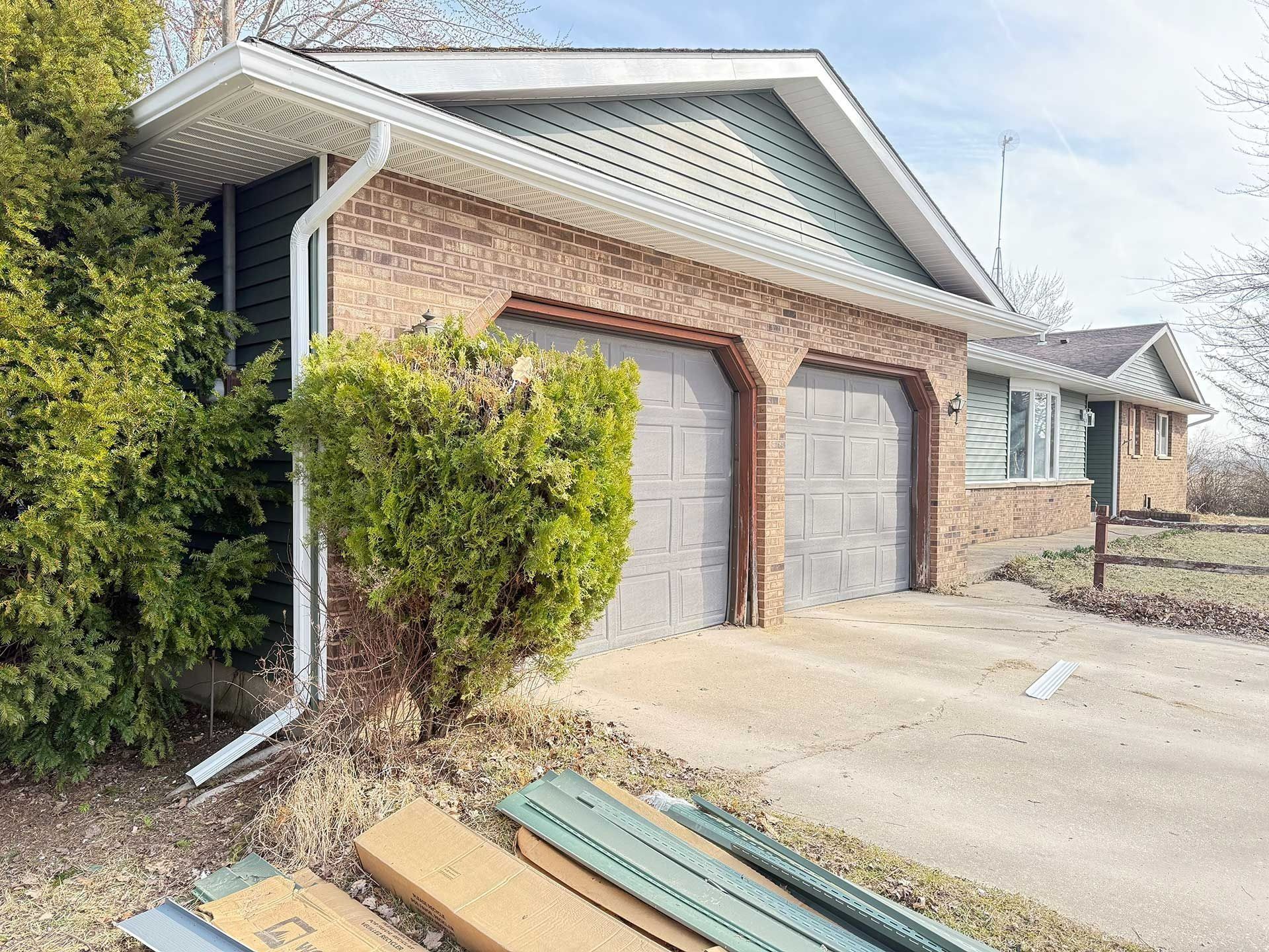 Brick garage with two gray doors, green shrub, and siding boards stacked on the driveway