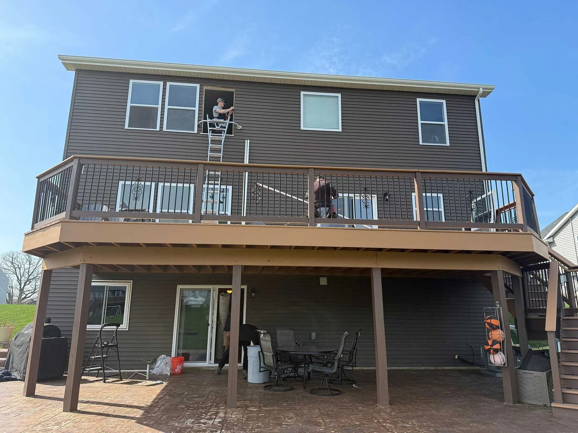 Two-story brown house with a deck and people on the balcony, viewed from the backyard.