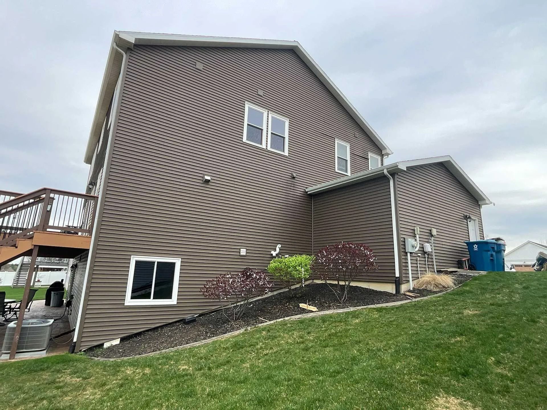 House with wavy brown siding on a grassy slope, with a deck and cloudy sky