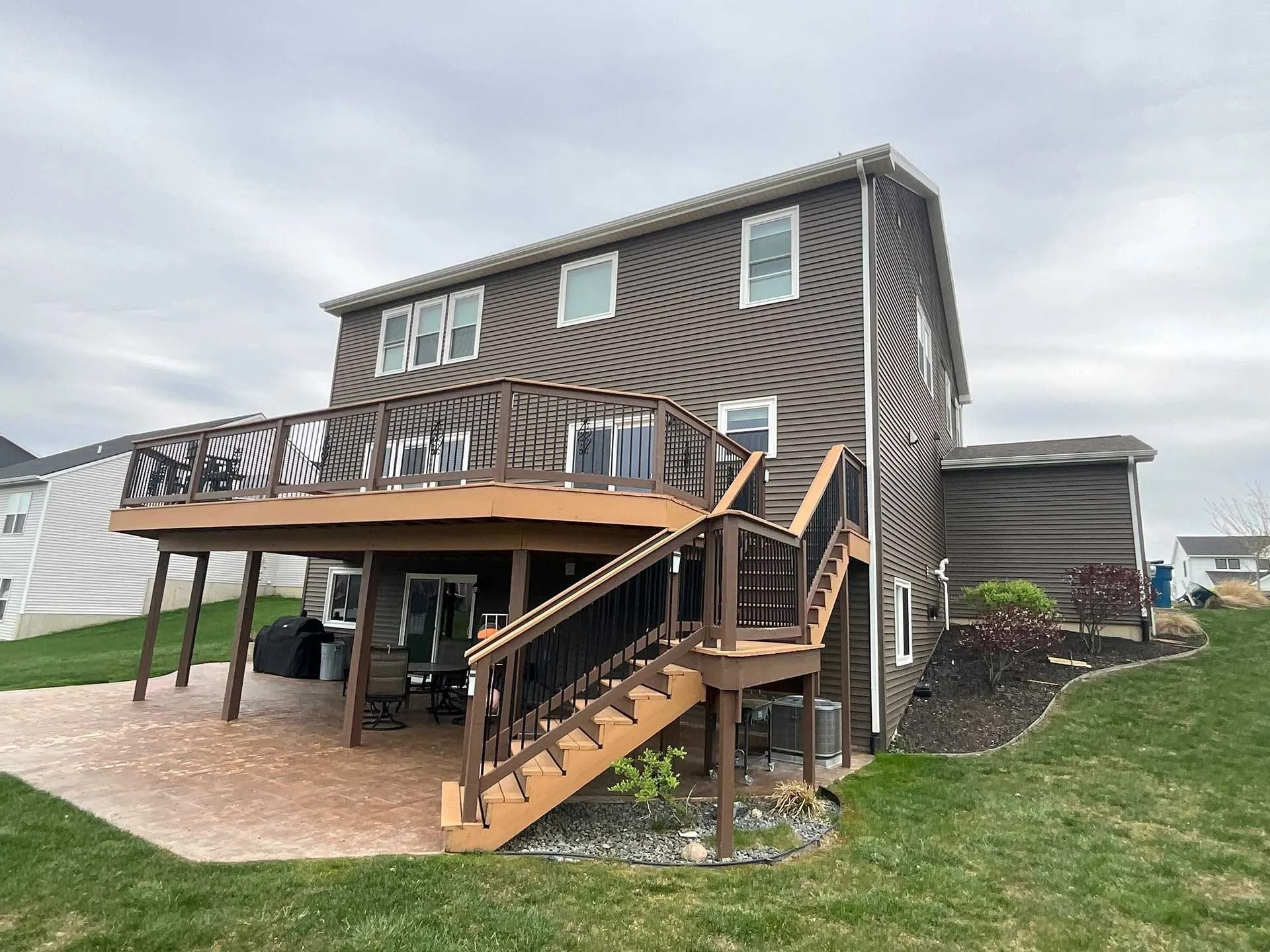 Two-story brown house with wooden decks and exterior stairs on a grassy lot under cloudy sky