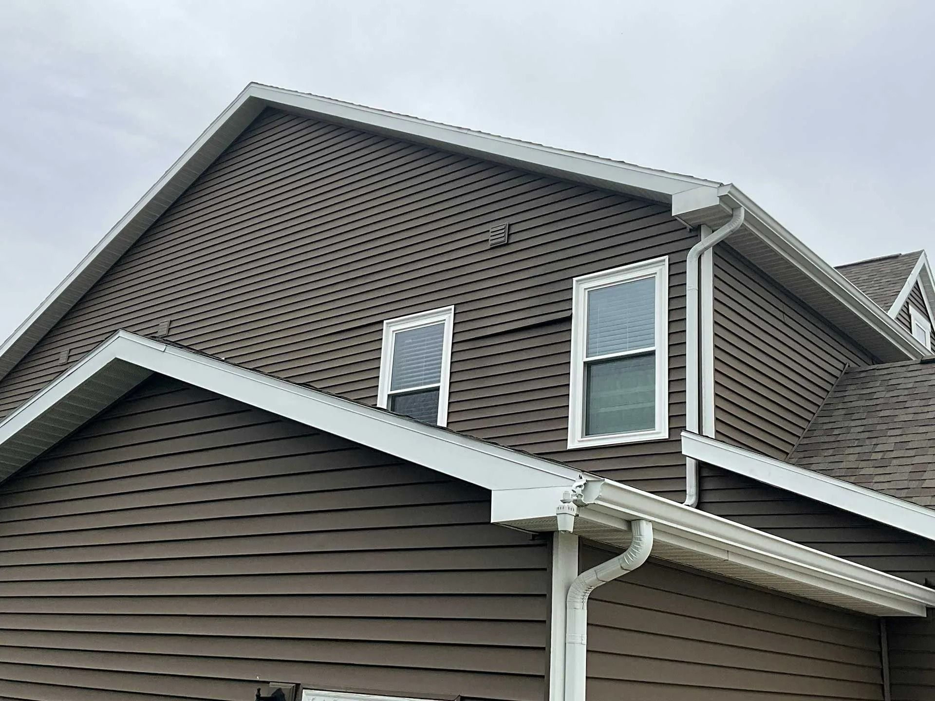 Brown house exterior with white trim and gabled roof, showing two upper windows and a gutter downspout