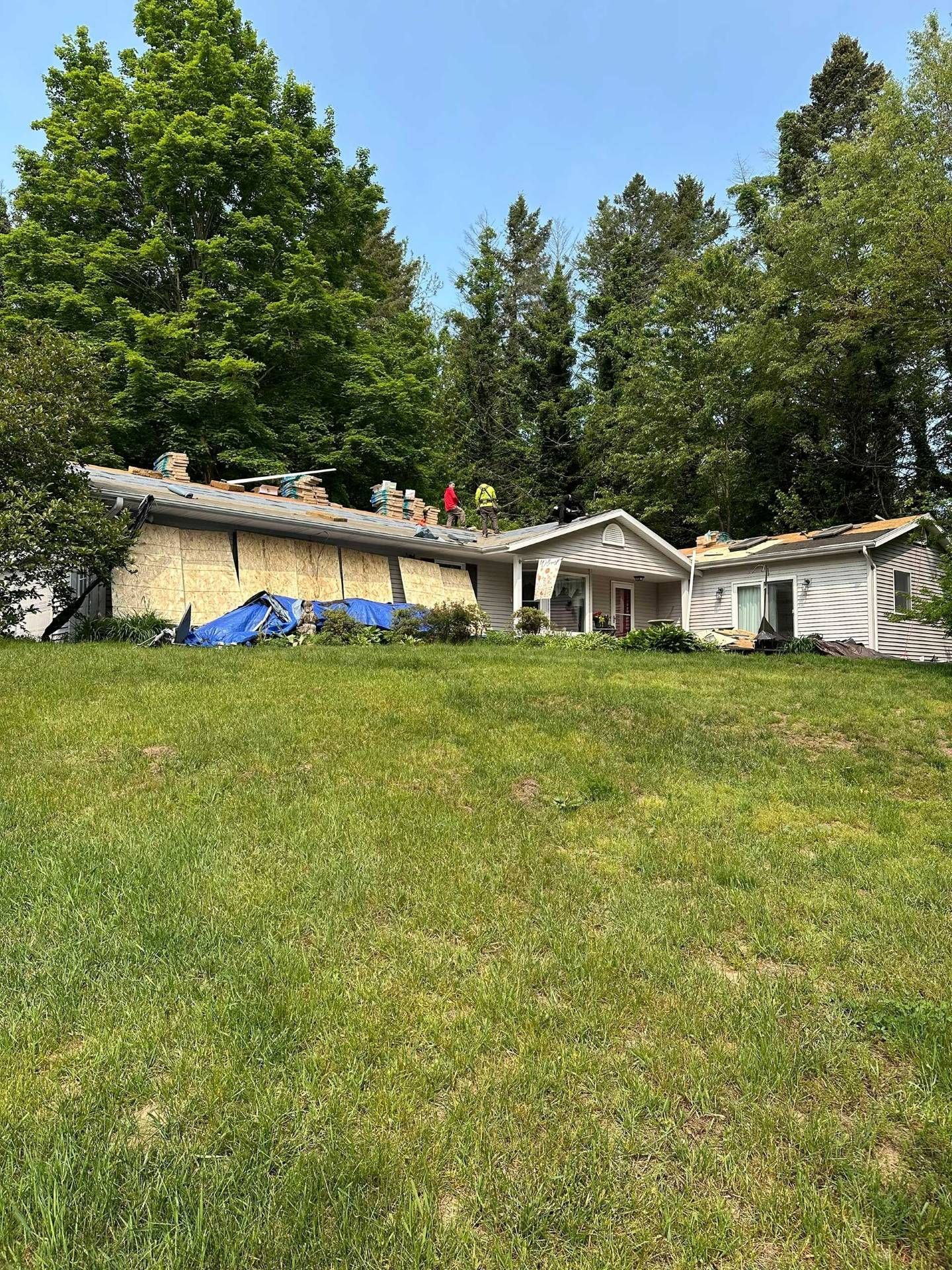 Wooded hillside with a small house, grassy slope, and blue tarp under a clear sky