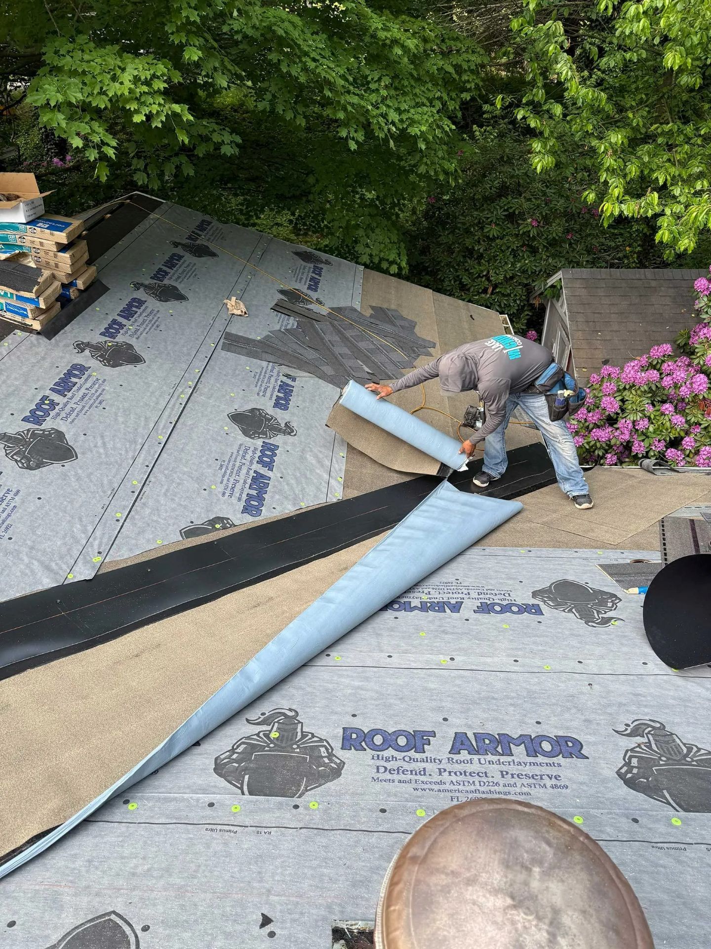 Worker installing roofing underlayment on a sloped roof near pink flowers and green trees