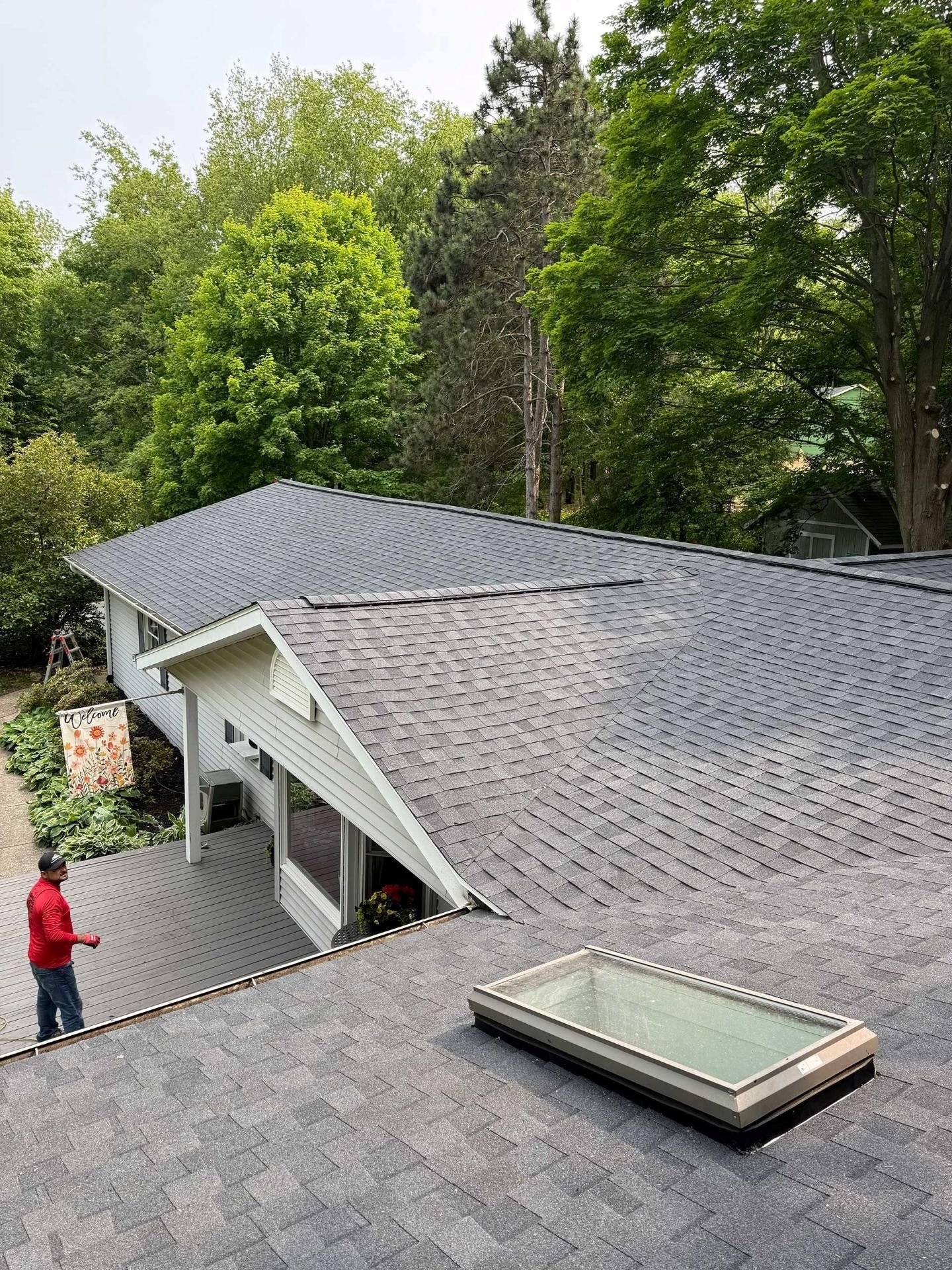 Roof with new gray shingles, skylights, and a worker on a sloped roof beside trees