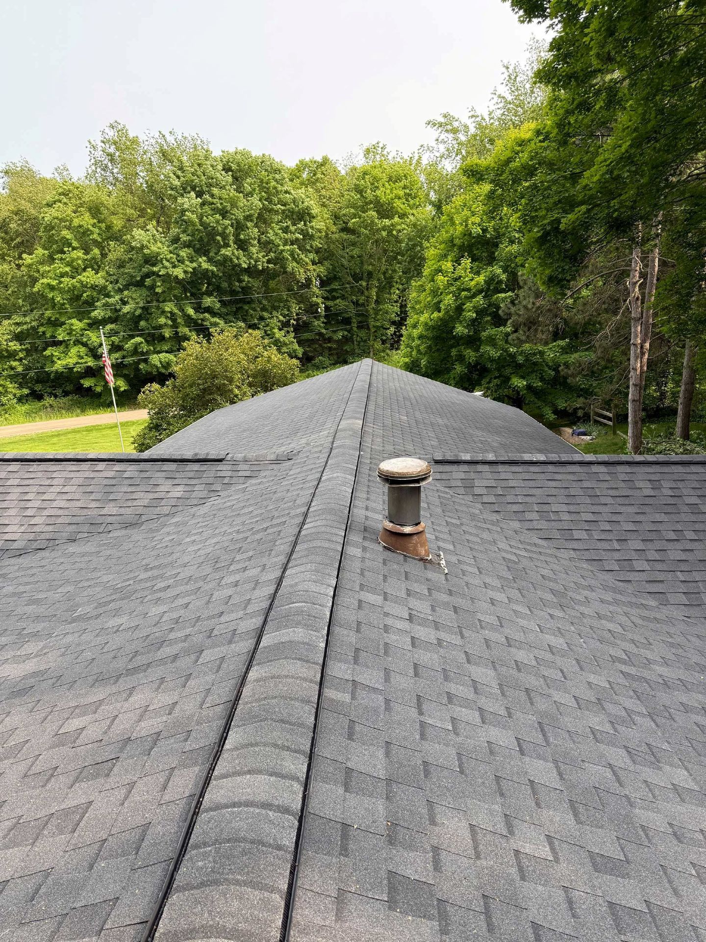 Roof shingles with a vent pipe, viewed toward a tree-lined yard under a bright sky