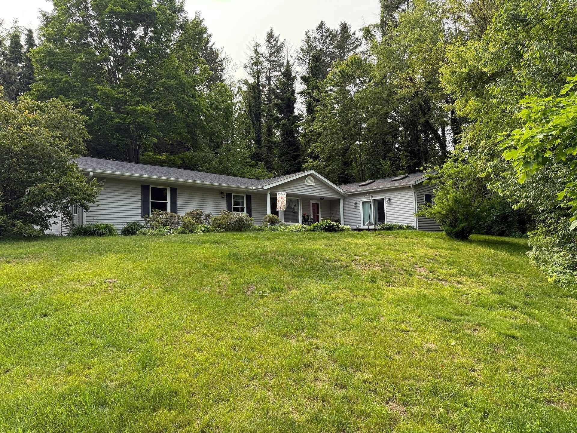 Single-story house with a gray roof on a grassy hillside, surrounded by dense green trees.