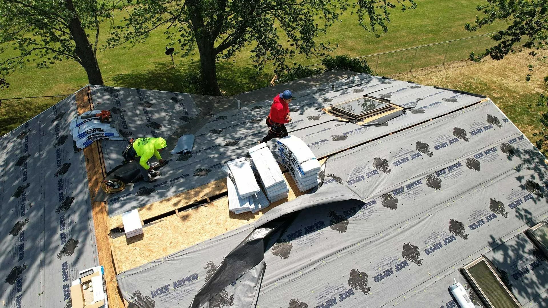 Workers installing roof shingles on a house, with gray underlayment and stacks of materials on the roof.