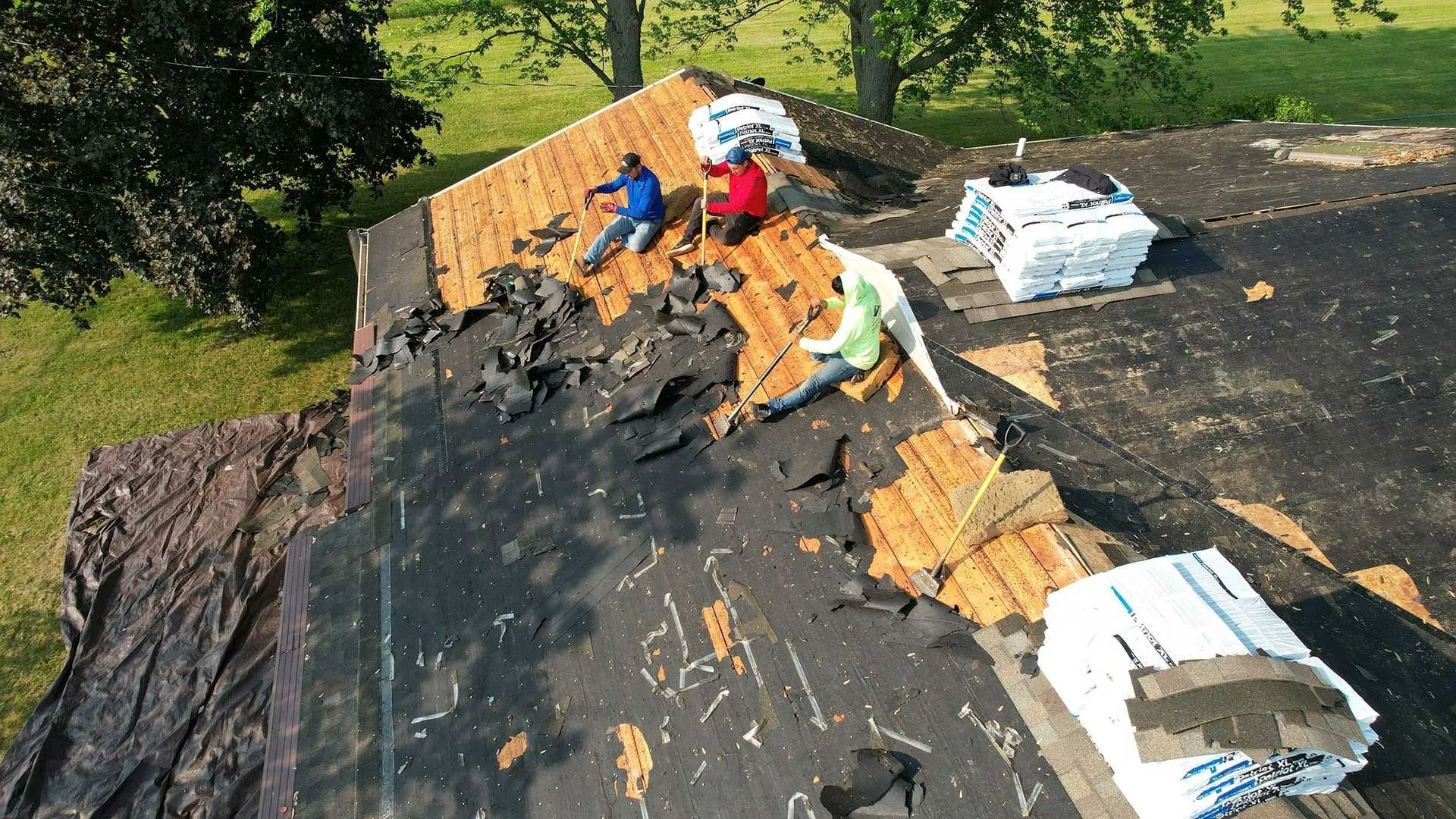 Workers repairing a shingled roof with black underlayment and stacked materials on top