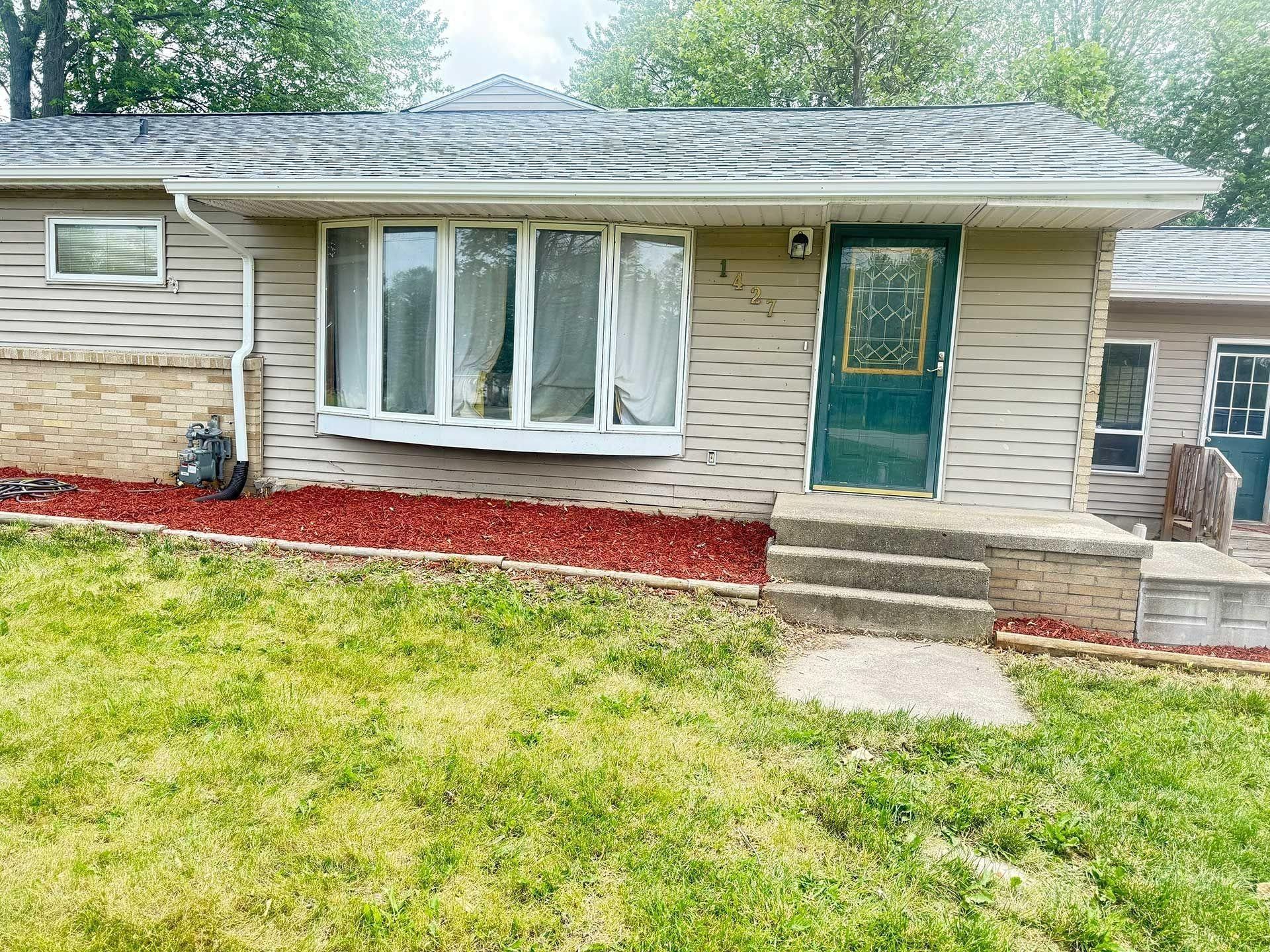 Front of a beige house with a green door, bay window, small porch, and red mulch beds.