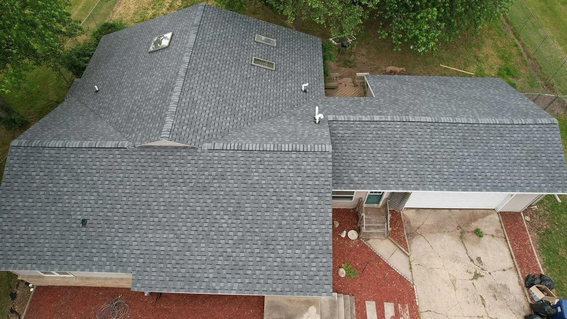 Aerial view of a house with dark gray shingle roofs and a small covered patio beside a wooded yard.
