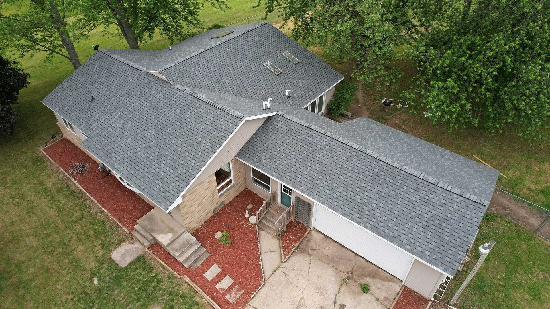 Aerial view of a gray-roofed suburban house with a red brick entry and white garage, surrounded by trees