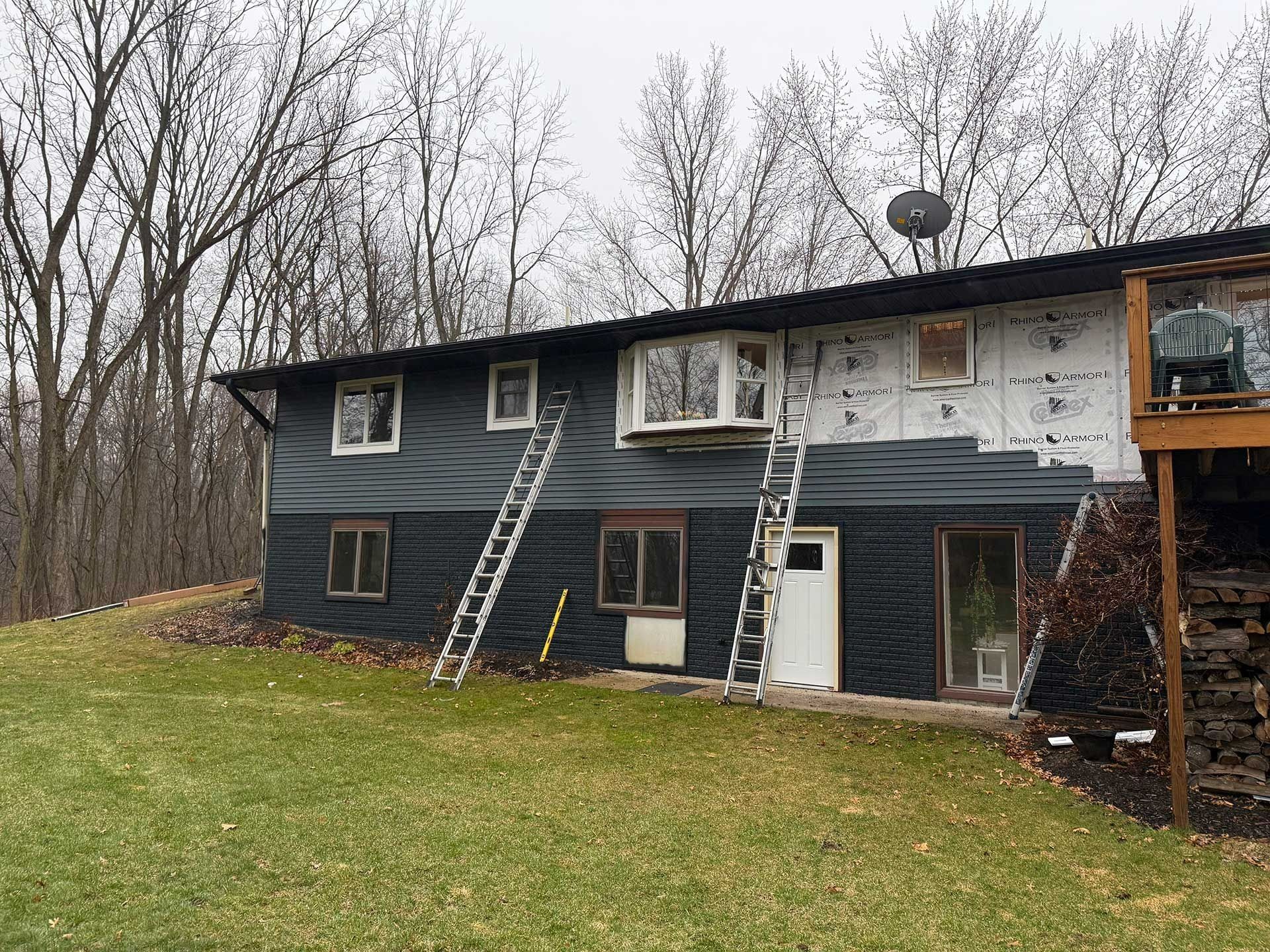 Two-story house with damaged siding and a ladder leaning against the roof, in a wooded yard.
