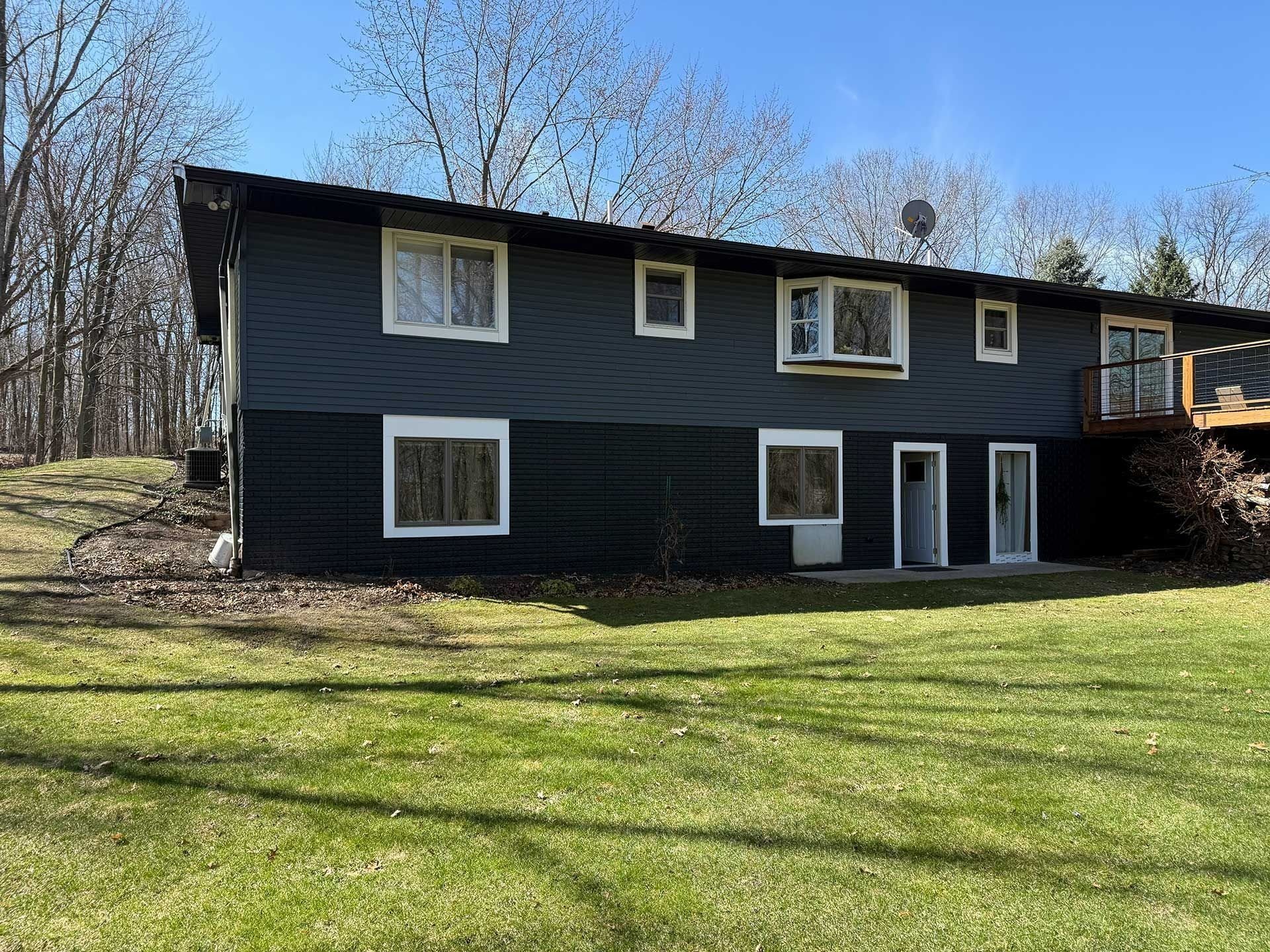 Dark two-story house with white-trim windows on a grassy slope near bare trees.