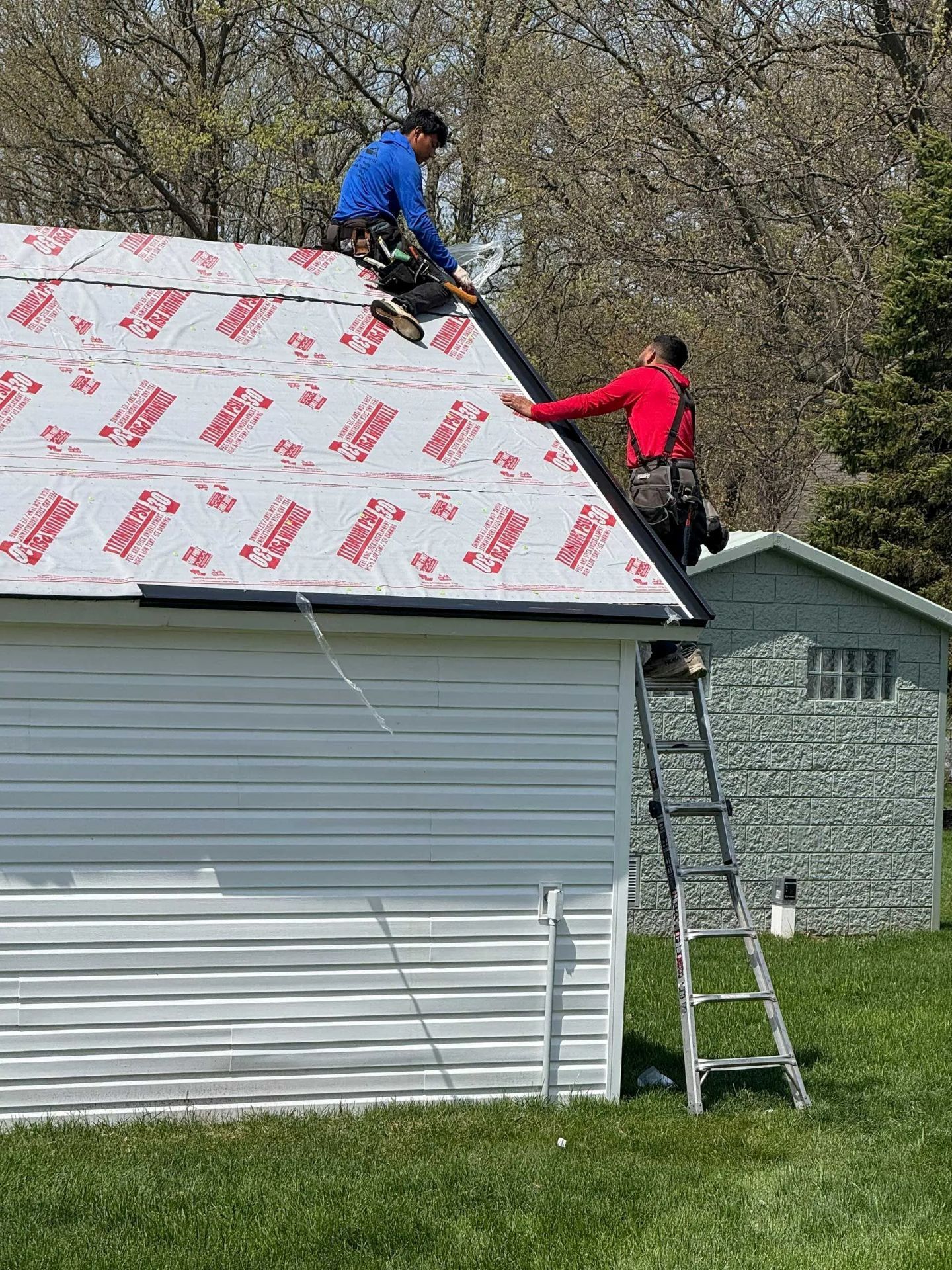 Two workers installing roofing underlayment on a house, with one on the roof and one on a ladder.