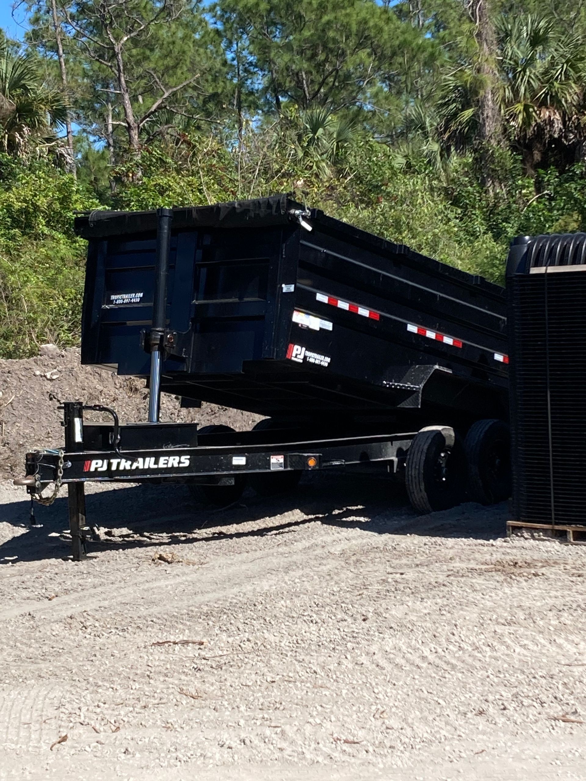 Black dump trailer tilted on a gravel lot, with trees and a hillside in the background