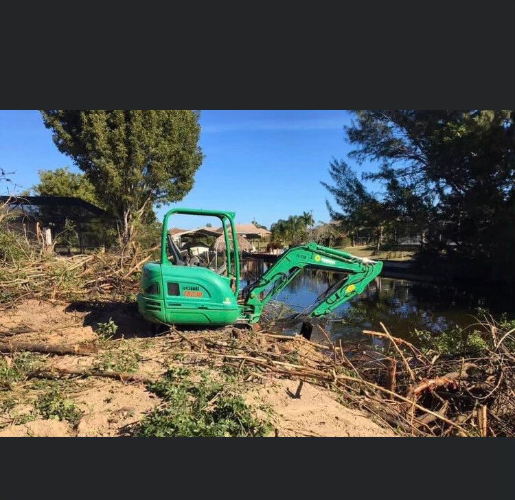 Green excavator by a muddy canal, its arm lowered amid brush and trees in a sunny rural area