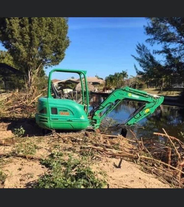 Green mini excavator working beside a creek in a wooded area under a clear blue sky