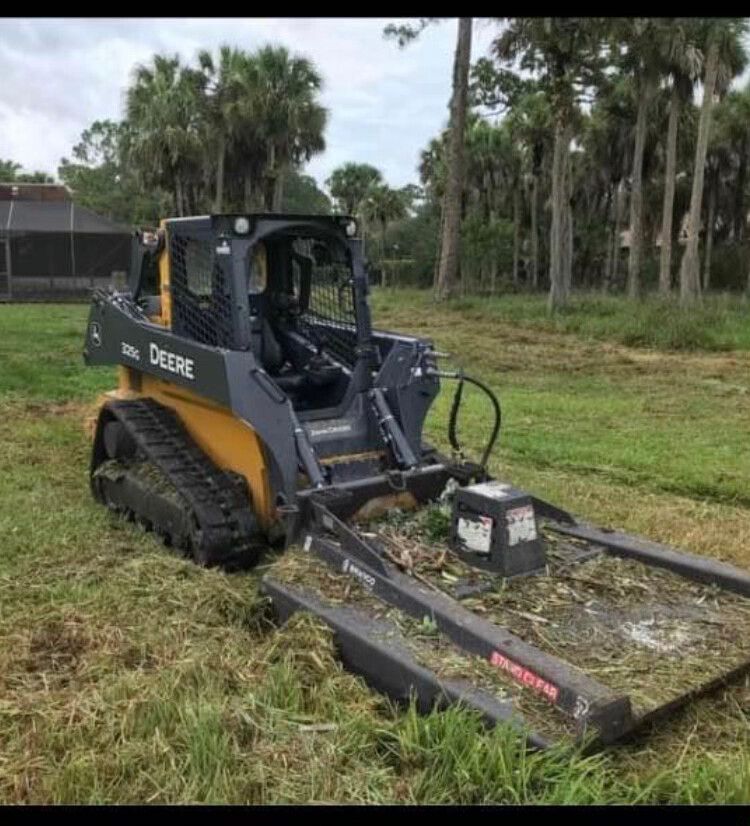 John Deere skid steer with mower attachment cutting grass in a palm-lined field.