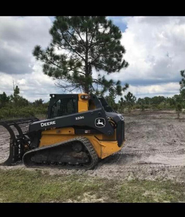 Yellow and black John Deere skid steer on muddy ground near a tall pine tree under cloudy sky
