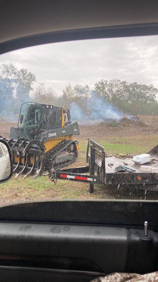 Black excavator in a grassy field with smoke rising, viewed from inside a vehicle.