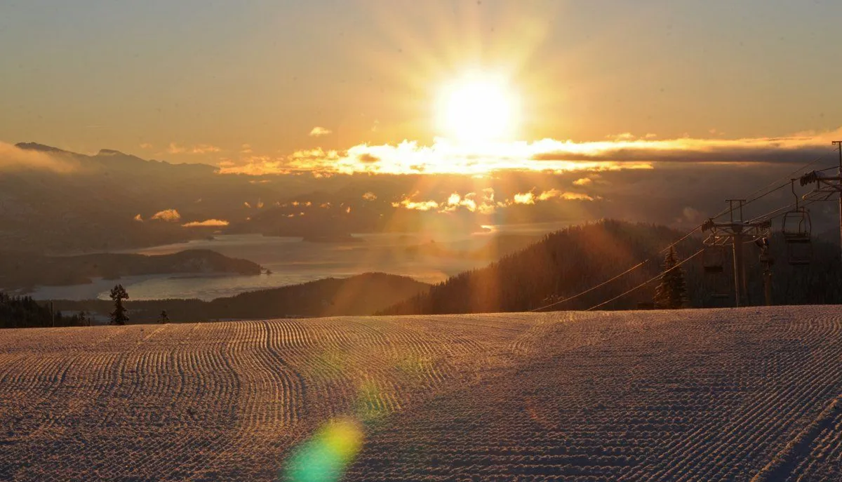Sunset over snowy mountains, with bright sun, clouds, and ski slope in the foreground