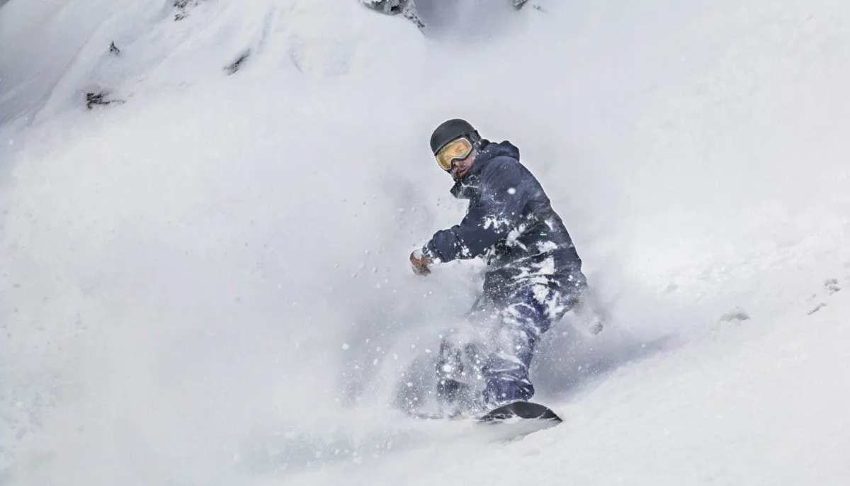 Snowboarder carving through deep powder on a snowy slope, wearing dark winter gear and goggles