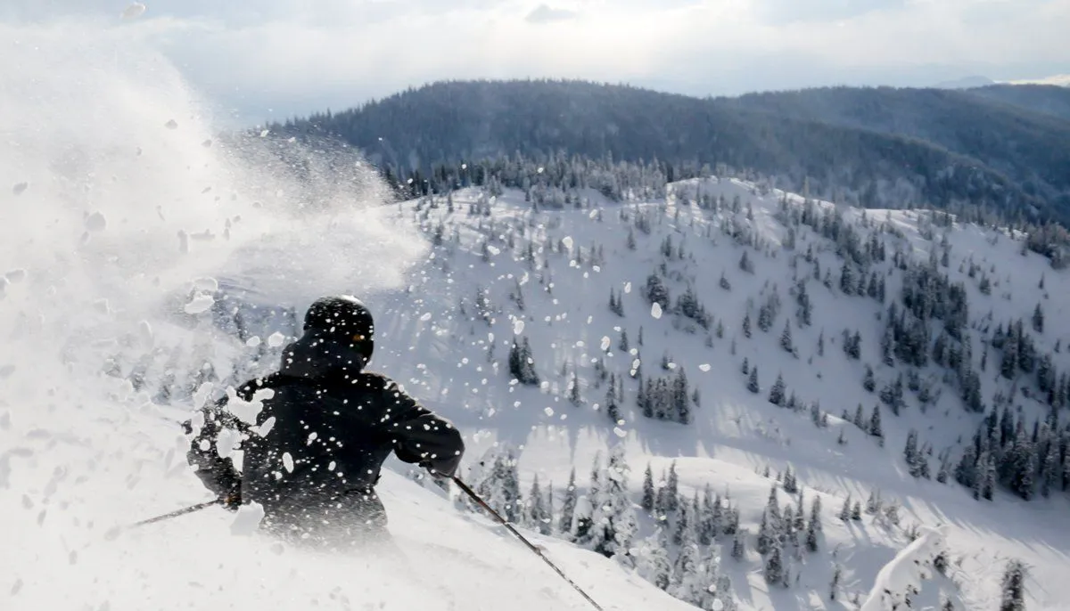 Skier carving through fresh powder on a snowy mountain slope with forested ridges in the background