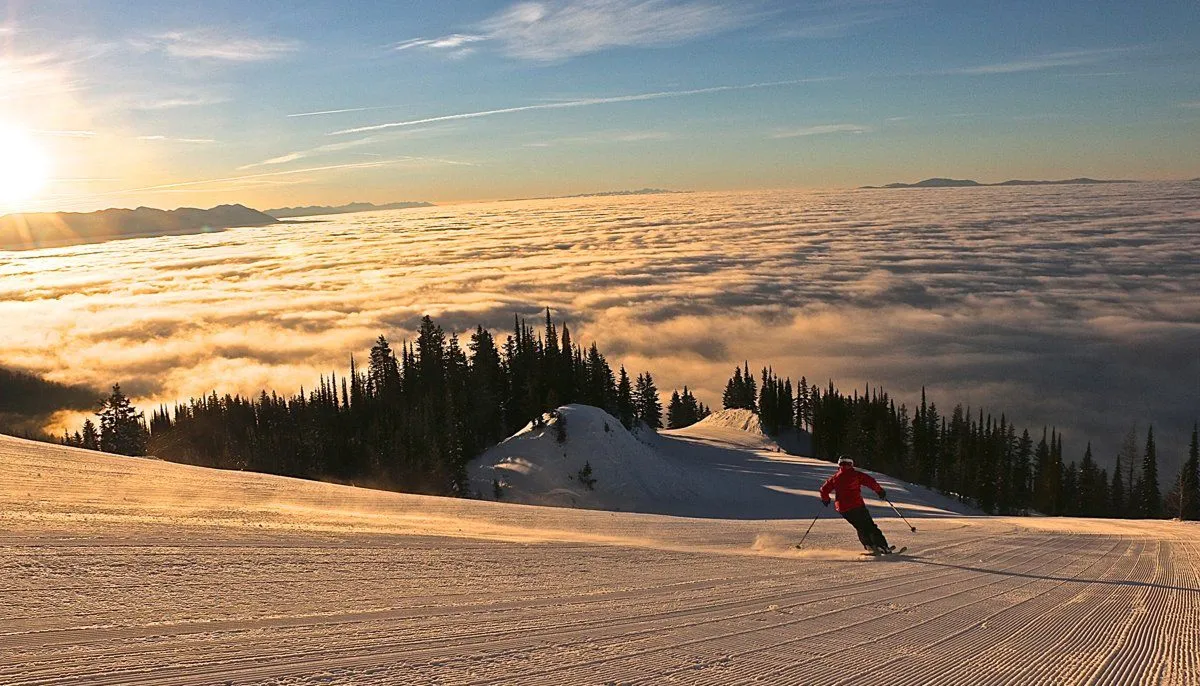 Skier in red carving down a snowy slope above a sea of clouds at sunrise