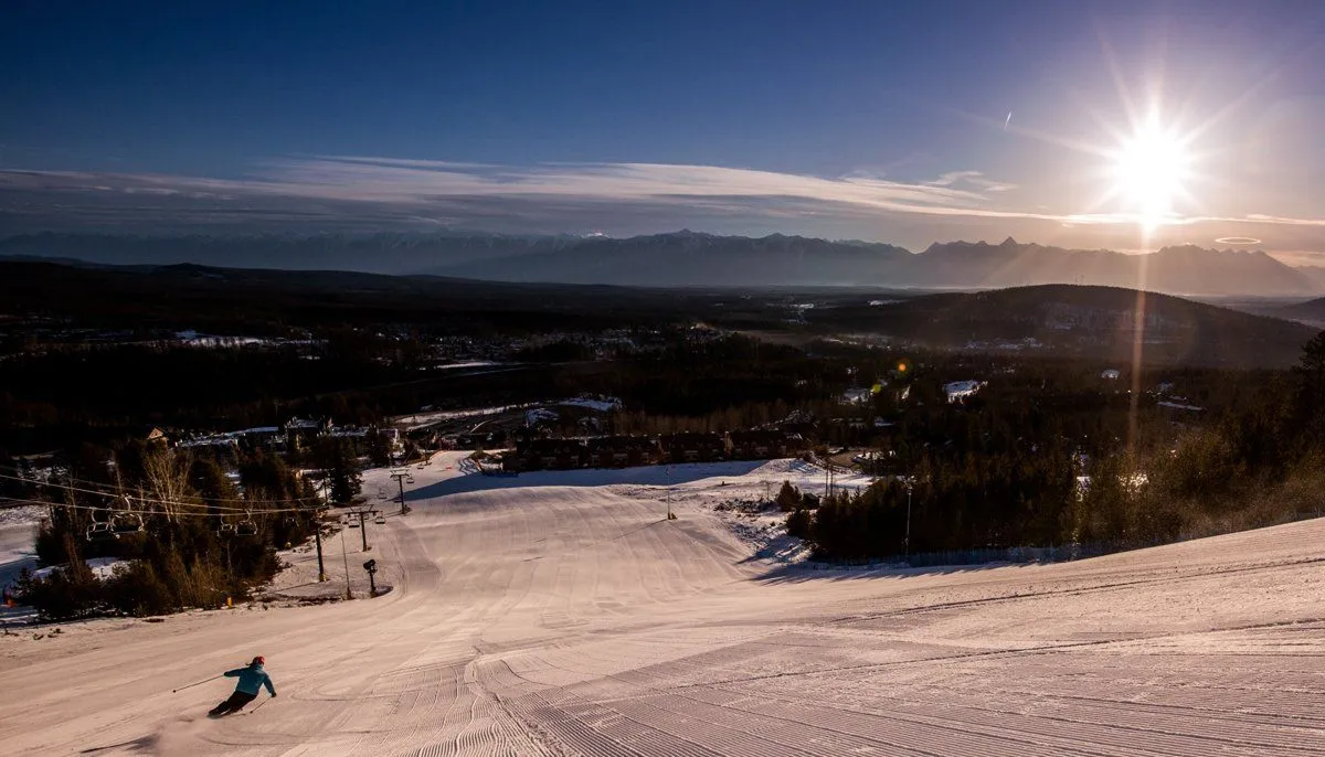 Snowy ski slope at sunset with a lone skier descending and a bright sun over distant hills
