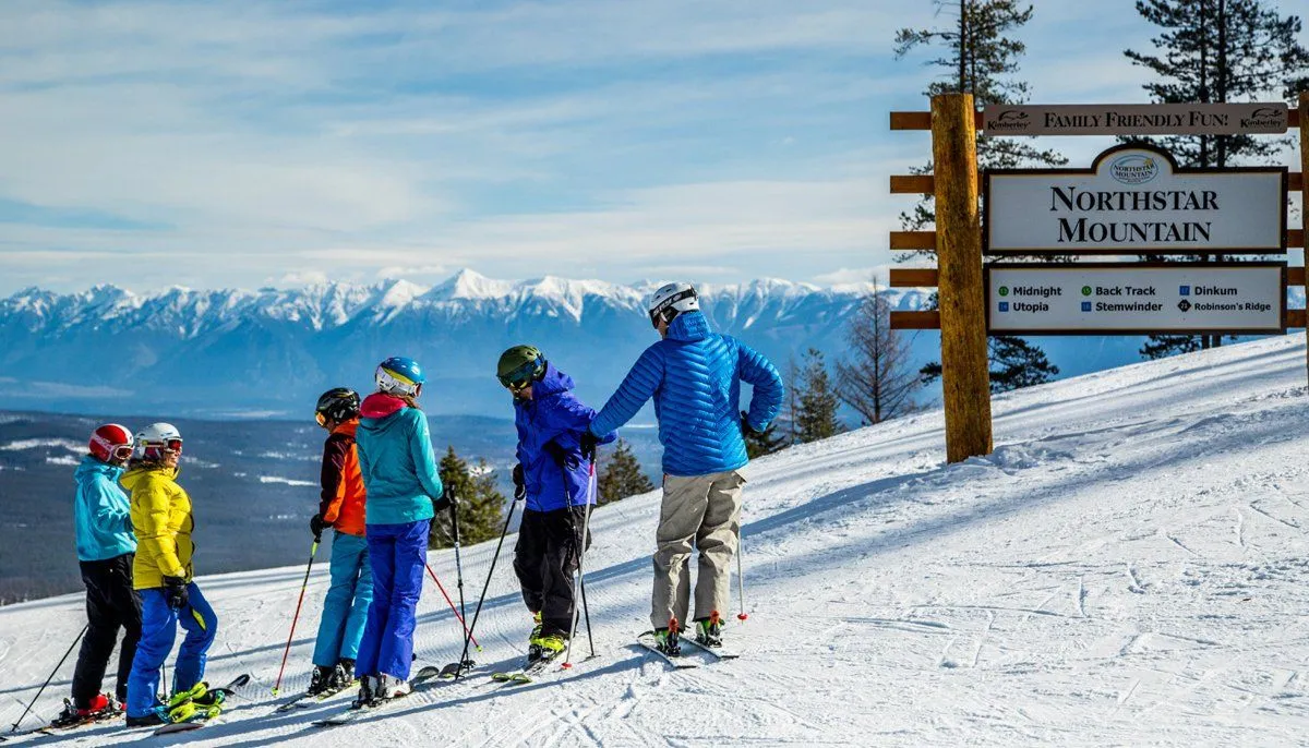 Skier in orange pants lands a jump on a snowy slope with mountain valley and chairlifts behind.