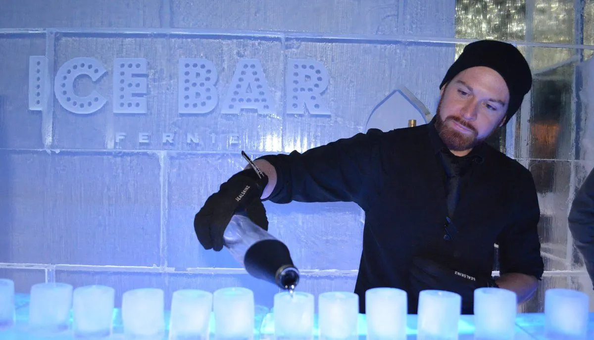 Bartender pouring a drink on an ice bar, with ice cups lined up and “ICE BAR” sign in the background