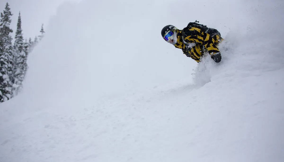 Snowmobiler in yellow gear airborne over a snowy slope, with trees in the background