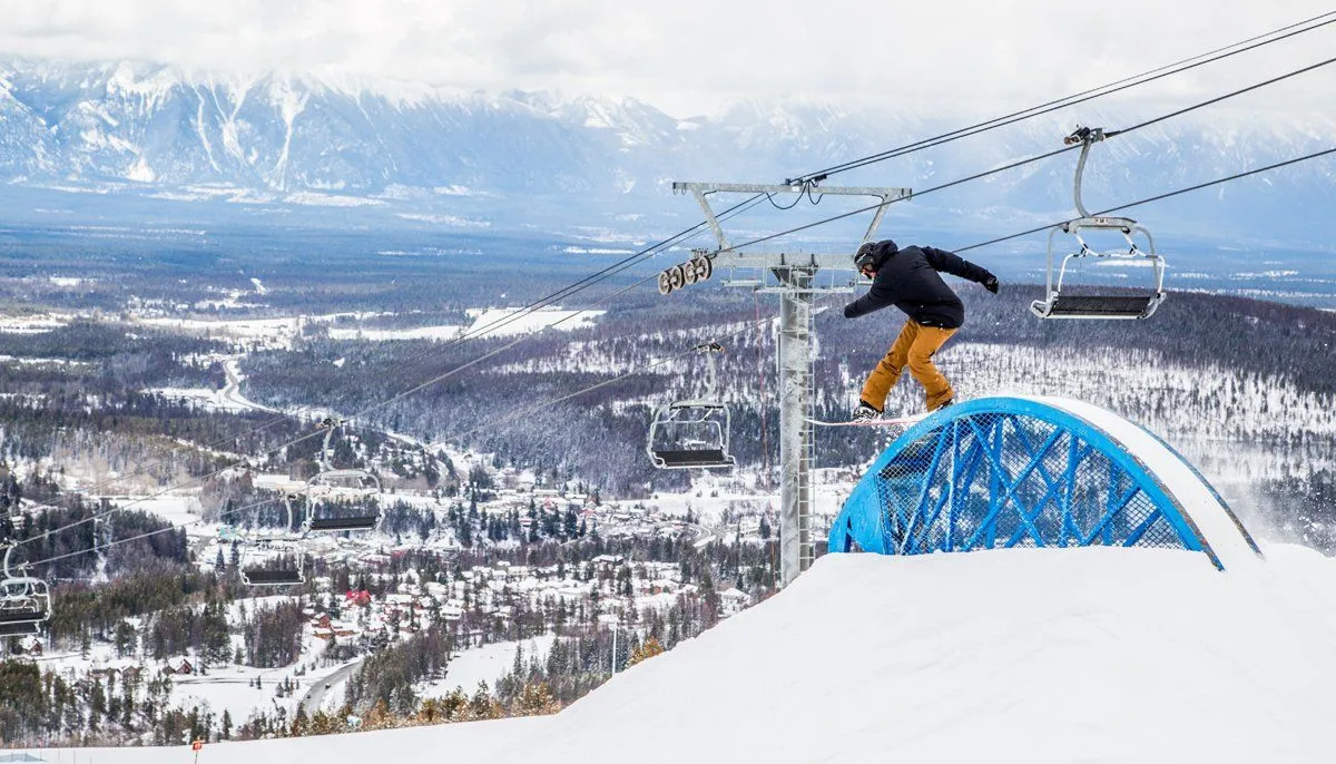 Snowboarder mid-jump on a blue rail above a snowy mountain ski slope with chairlifts in the background