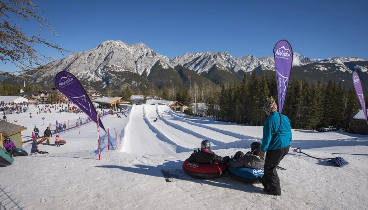 People sledding on a snowy mountain slope with pine trees and jagged peaks in the background.