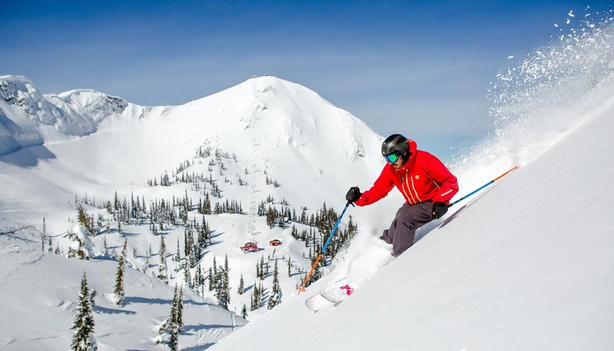 Skier in red jacket carving down a snowy mountain with snow spraying, alpine peaks in the background