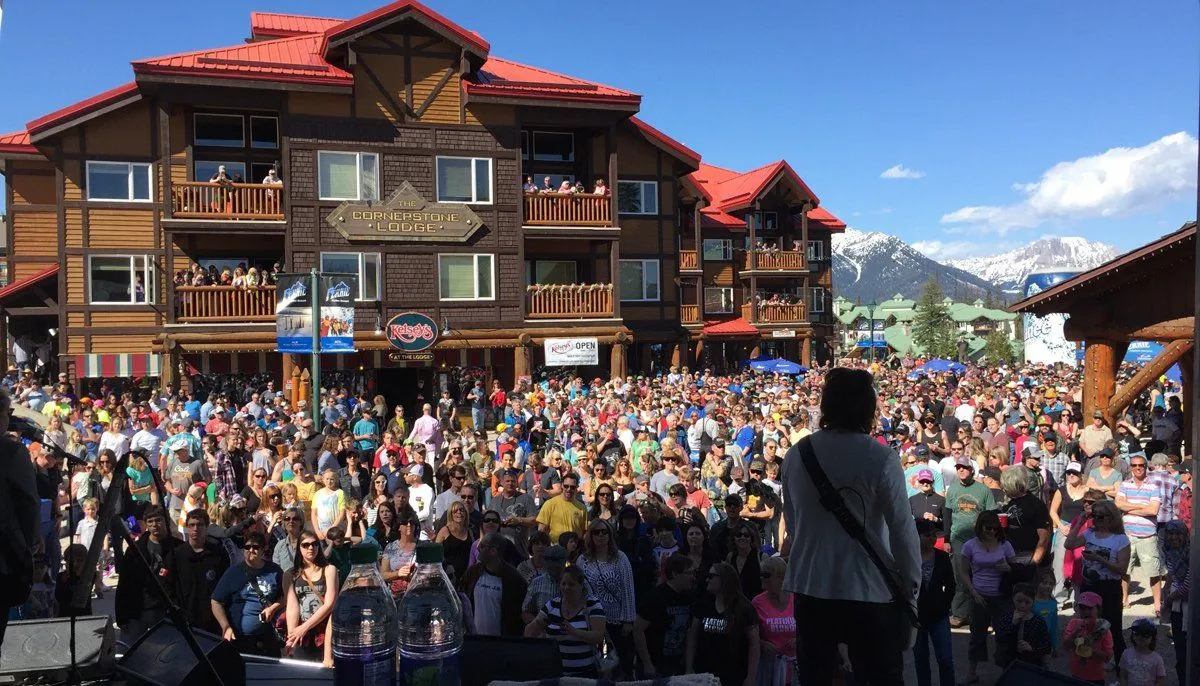 Large crowd gathered before a chalet-style building in a snowy mountain resort, seen from a stage.